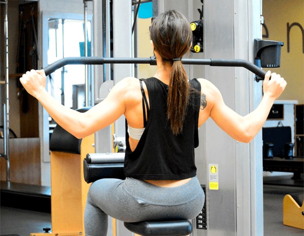 A person with long hair in a ponytail is using a lat pulldown machine at the gym. They are facing away from the camera, wearing a black sleeveless shirt and gray leggings. The gym equipment and surroundings, representative of affordable gym memberships, are visible in the background.