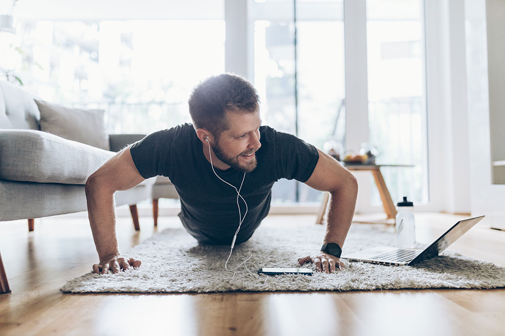 A man is doing push-ups on a rug in a bright living room, engaging in a healthy workout while looking at a laptop. He is wearing a black t-shirt, dark pants, and earphones. Nearby, a smartphone and water bottle lie on the floor as natural light streams through large windows in the background.