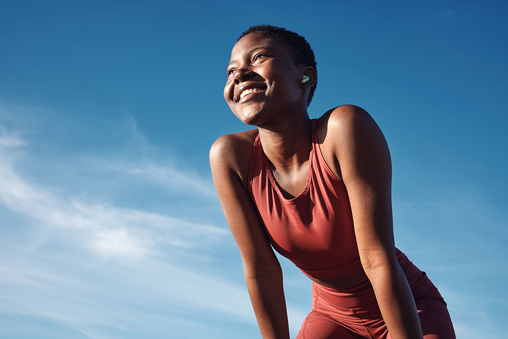 A person wearing a red athletic outfit is smiling and bending slightly forward against a bright blue sky with light clouds in the background. The individual appears to be outdoors, enjoying a sunny day, showcasing their fitness and healthy lifestyle possibly supported by a gym membership.