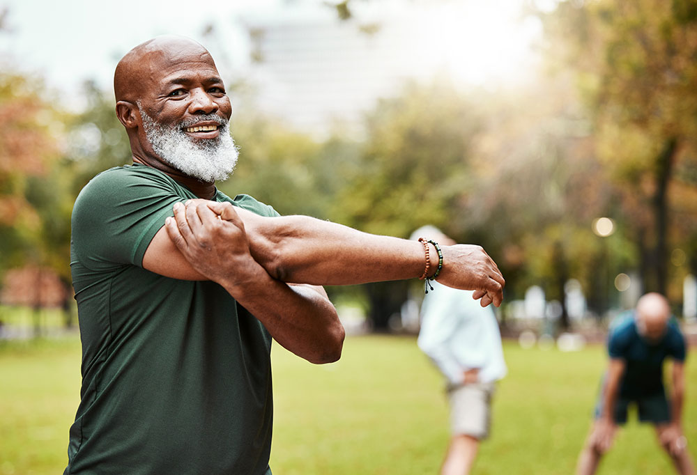 A smiling elderly man with a white beard stretches his arms in a park. Wearing a dark green shirt, he appears to be enjoying a group exercise session focused on fitness and healthy living, with others stretching in the background amidst lush greenery.