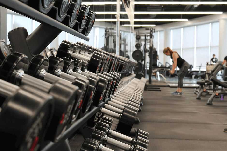 A gym with a row of black dumbbells on a rack in the foreground. In the background, a person with blonde hair wearing a black tank top bends over while lifting weights. Several other exercise machines and equipment are visible, providing everything needed for an effective beginner gym workout. The room is well-lit.