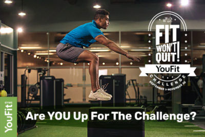 A man in athletic wear is mid-air during a box jump at a gym. Behind him, exercise equipment is visible. The text reads, "Are YOU Up For The Challenge?" along with a graphic stating, "Fit Won't Quit Check-in Challenge" and the YouFit logo on the left side.