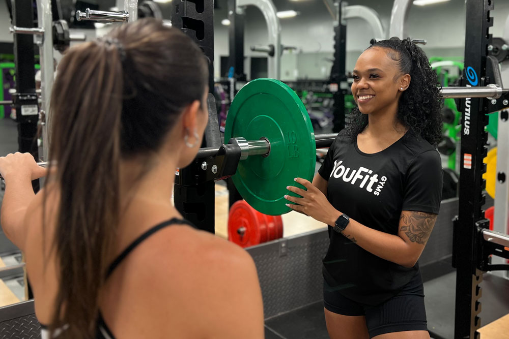 Two people at a gym: one preparing to lift weights, facing away from the camera, and another, wearing a "YouFit" shirt, is assisting by placing a green weight plate on the barbell. The gym, vibrant with various fitness equipment in the background, offers an inviting environment for any workout.