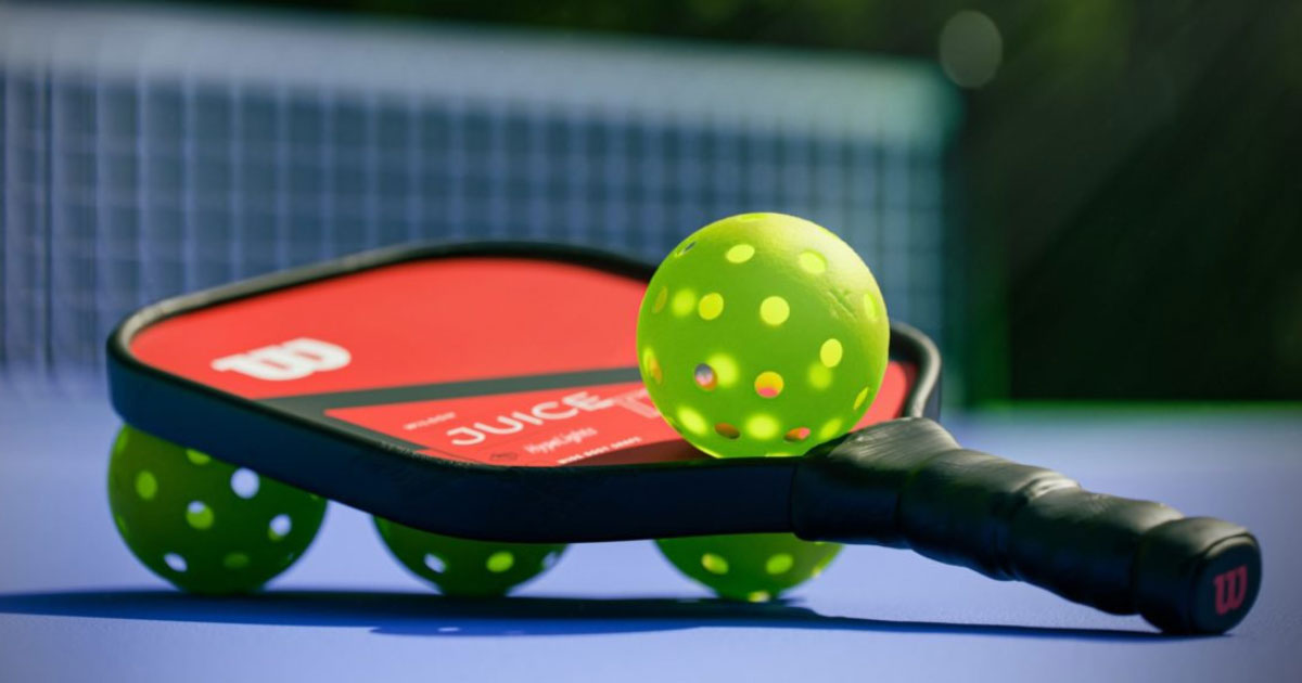 A pickleball paddle rests on a blue court near a net, with bright green pickleballs placed around and on top of the paddle. The paddle has a black handle and a red face with white text. The background is partially blurred but shows the pickleball net—a perfect setup for your next fitness workout.