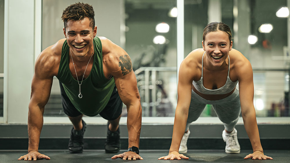 Two people are in a gym performing push-ups side by side. The person on the left, wearing a green tank top, has a muscular build and a tattoo. The person on the right, wearing a gray sports bra, has a fit appearance. Both are smiling, clearly enjoying their workout and making the most of their gym membership.