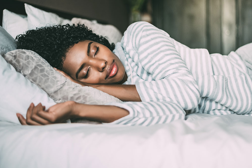 A person with curly hair peacefully sleeps on their side in a bed, wearing a striped pajama top and resting their head on a patterned pillow. A white blanket covers part of their body, creating a cozy and serene scene— the perfect rest after maintaining a healthy exercise routine.