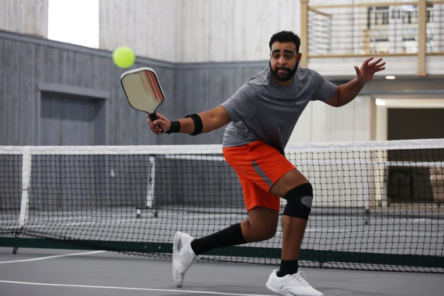 A man is playing pickleball on an indoor court, wearing a gray shirt and orange shorts. He swings his paddle to hit a green ball, focusing intensely near the net. The scene exudes fitness and energy, with a wooden railing visible in the background.