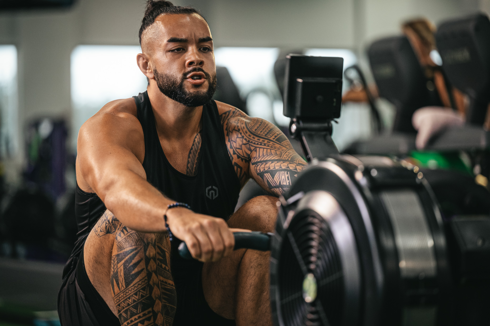 A man with tattoos works out on a rowing machine in a gym. Focused and determined, he wears a black tank top while pulling on the rowing handle. Surrounding exercise equipment highlights his dedication to fitness and maintaining a healthy lifestyle.