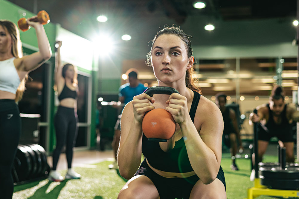 A woman is performing a squat exercise while holding an orange kettlebell in a gym. She is part of a small group training session, surrounded by others engaged in various exercises. The gym has bright lighting and green flooring designed for functional training.