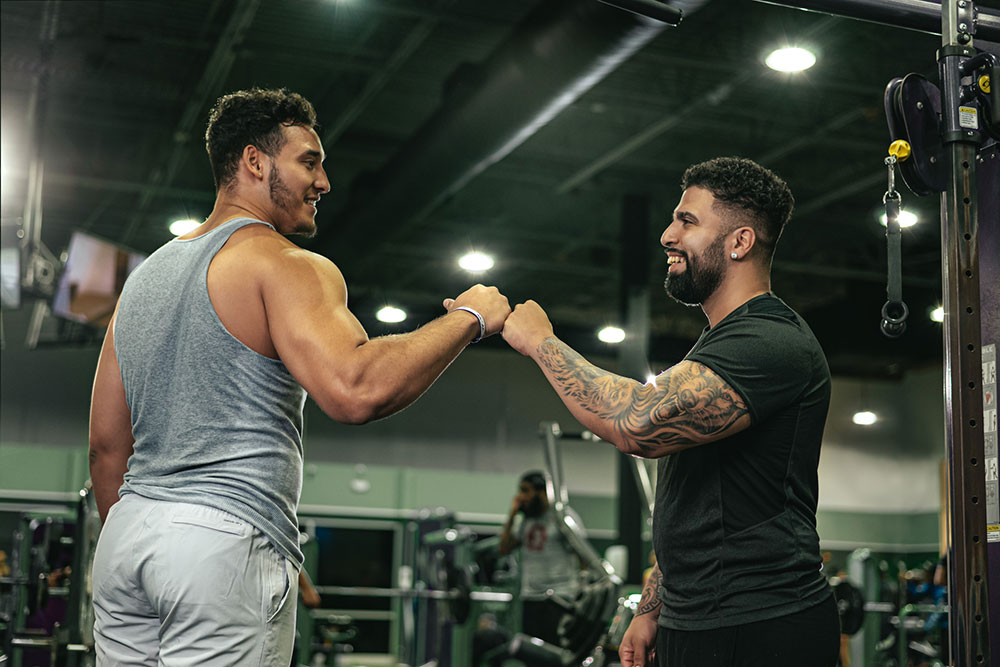 Two men are engaging in a fist bump at a gym, sharing a moment of camaraderie. The man on the left, wearing a gray tank top and white shorts, and the man on the right, in a black t-shirt with tattoos on his right arm, are both smiling. It's as if their silent "thank you" echoes amidst the gym equipment in the background.