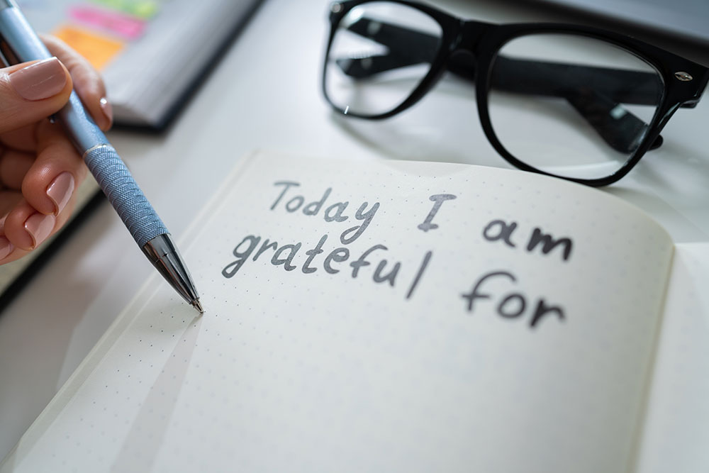 A hand holding a pen poised over an open notebook with dotted pages. The handwritten text on the page reads, "Today I am grateful for." Near the notebook, a pair of black glasses rests on the table, and colorful sticky notes featuring workout reminders are visible in the background.