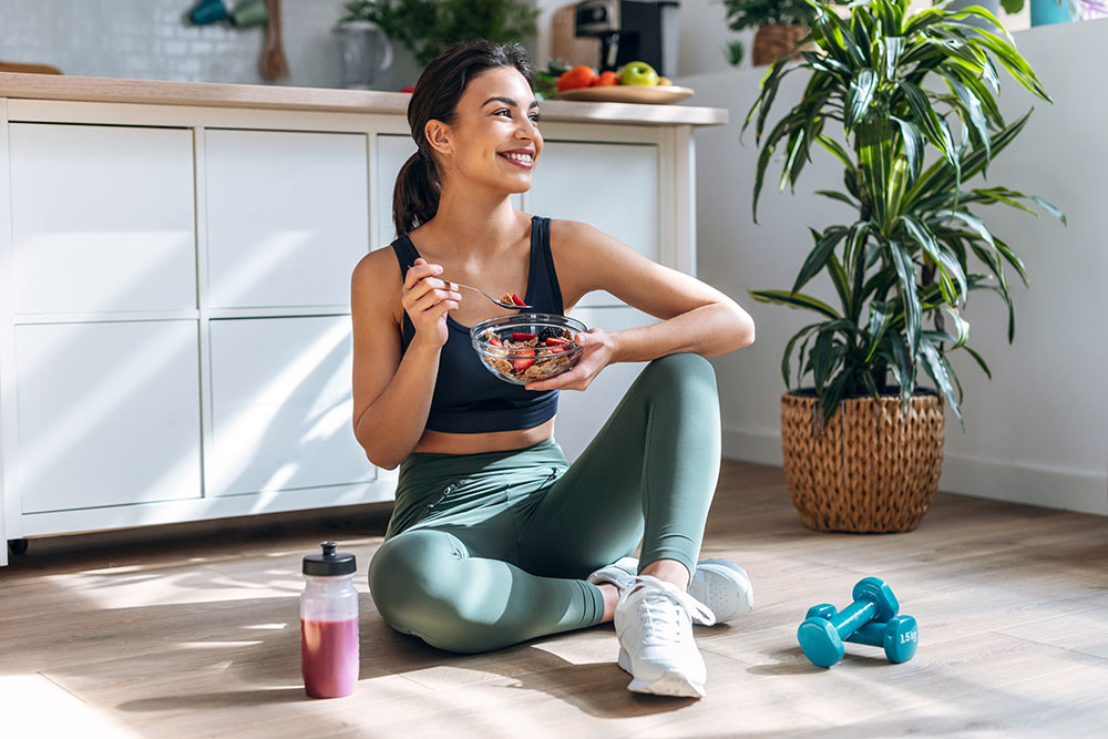 A woman in workout attire sits on the floor, holding a bowl of fruit salad, illustrating the question: should-you-eat-breakfast? She is smiling and appears relaxed. Around her, there are green dumbbells, a pink water bottle, and a potted plant, suggesting a home workout setting.