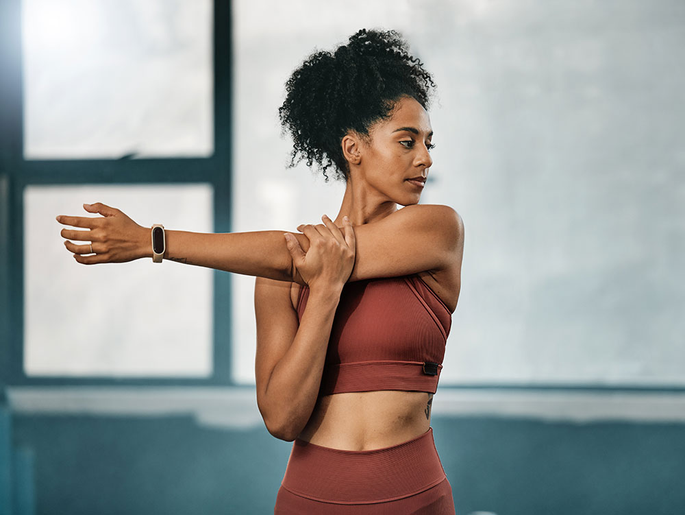 A woman wearing a matching red sports bra and leggings is standing indoors, stretching her arm across her chest. Embracing a healthy lifestyle, she has curly hair and is focused on her exercise. She sports a smartwatch on her wrist, with light streaming through large windows in the background.