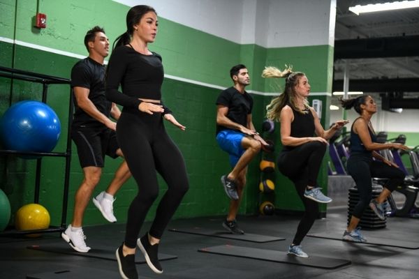 A group of five people in athletic wear are performing a jumping exercise in a gym. They are positioned on individual mats, and the background features green and white walls with various gym equipment like exercise balls and weights—perfect for classes for beginners.