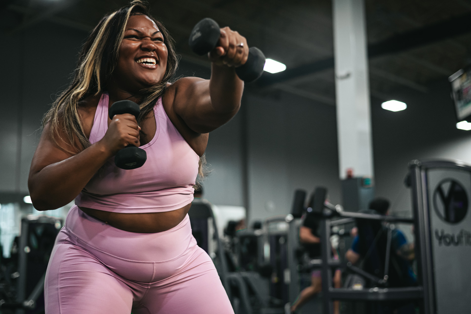 A woman wearing a pink sports bra and matching leggings is exercising with dumbbells in a gym, clearly making the most of her mental health day. She has a joyful expression on her face amidst several fitness machines and other people working out in the background.