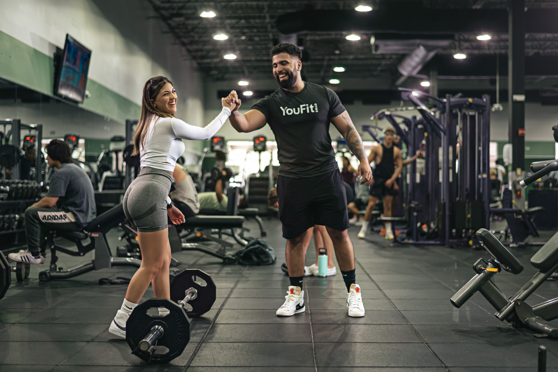 A man and a woman high-five each other in a gym. The woman, wearing a white top and gray shorts, stands next to a barbell on the floor, while the man, in a black "YouFit" t-shirt and shorts, smiles. Their camaraderie helps ease gym anxiety as other gym-goers and equipment are visible in the background.