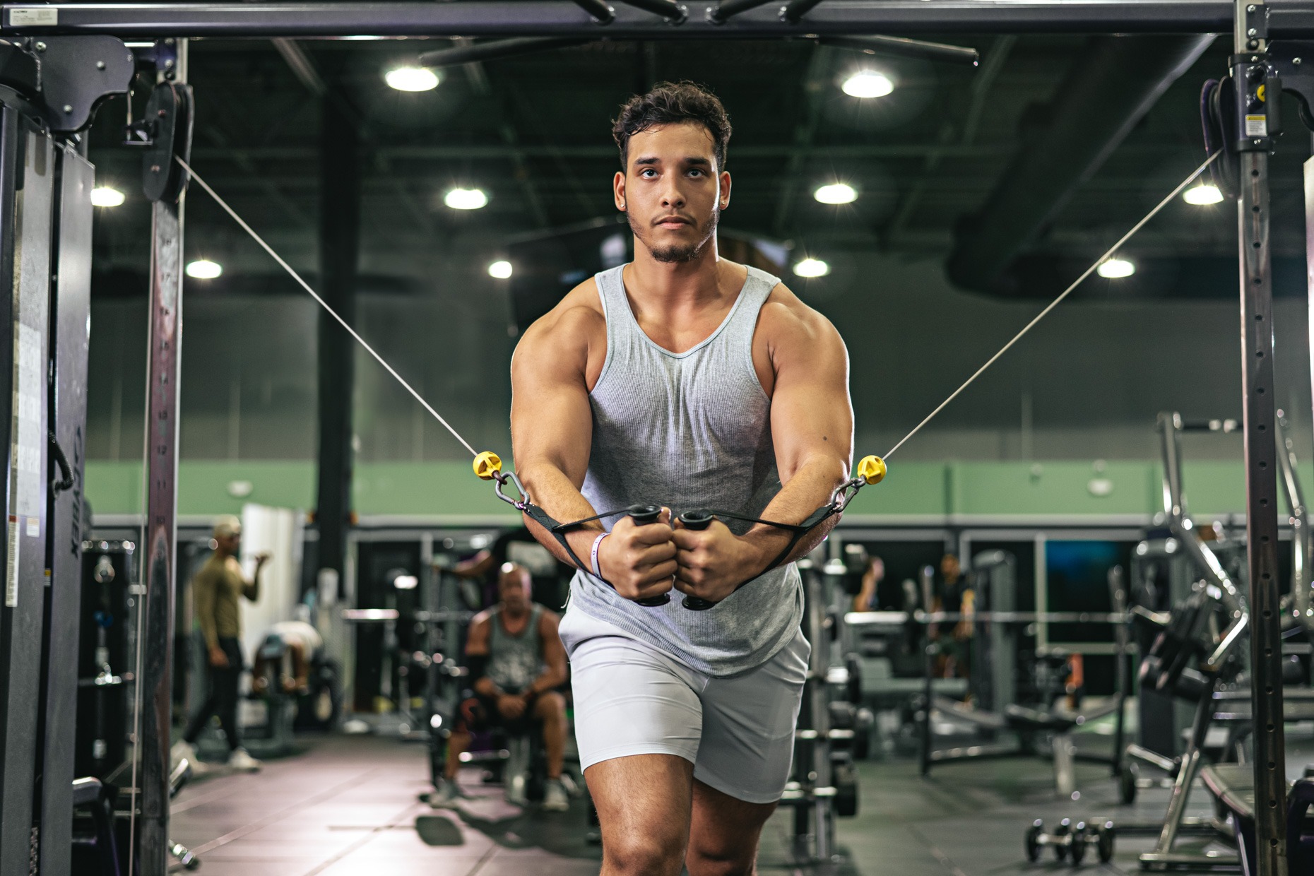 A man in a gray tank top and light-colored shorts is working out in a gym, using a cable machine for chest exercises. Other people and gym equipment are visible in the background. The lighting is bright, highlighting his focused expression and muscular build, showcasing his dedication to strength training and muscle growth.