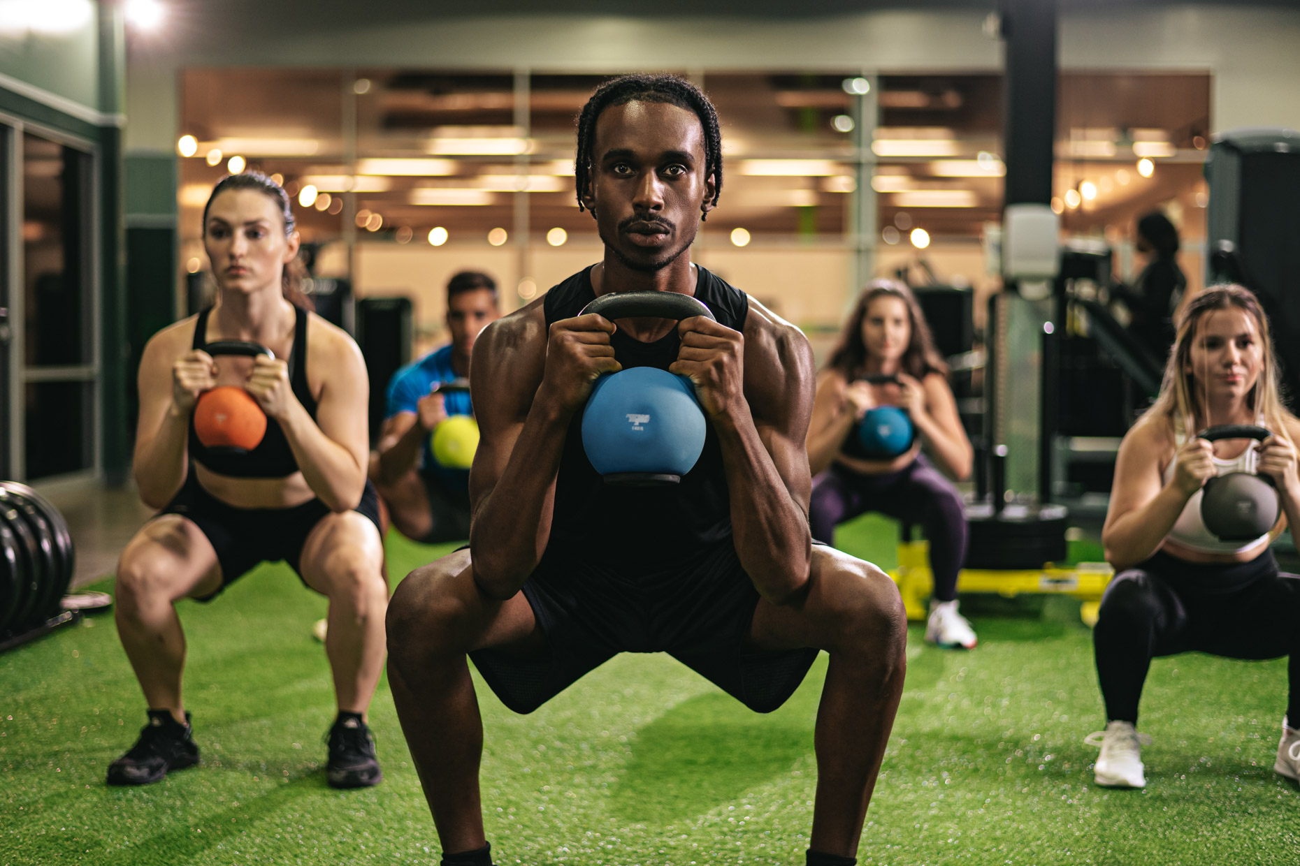 A group of five people performing goblet squats with kettlebells in a gym, as part of a HIIT session. A man in the front center holds a blue kettlebell. All participants appear focused and are positioned on a turf-covered floor with gym equipment in the background.