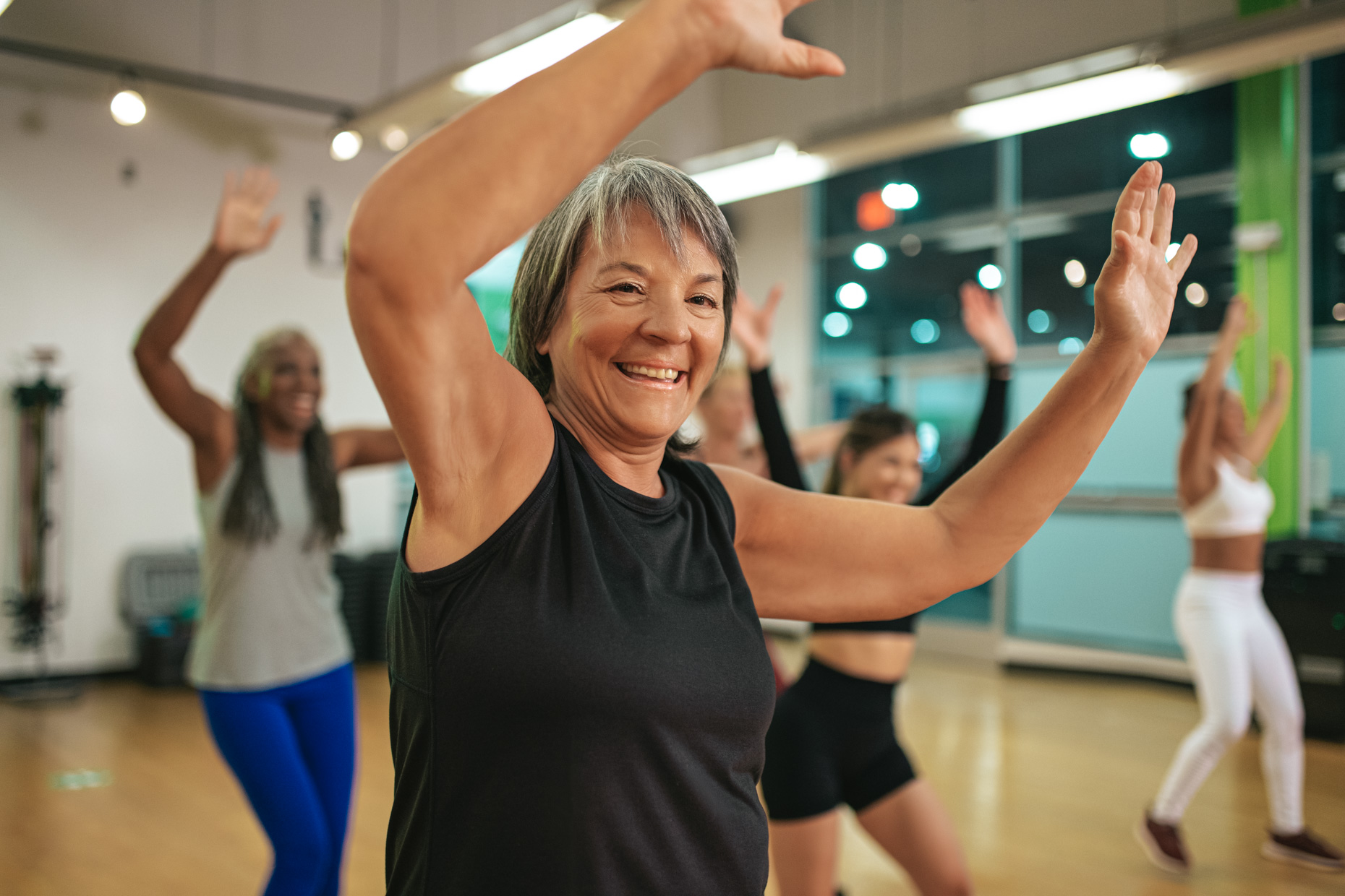 A group of enthusiastic women participate in a lively dance class in a spacious, bright room with large windows. A woman in the foreground, wearing a black sleeveless top, smiles widely with her arms raised, leading the functional and energetic movements.