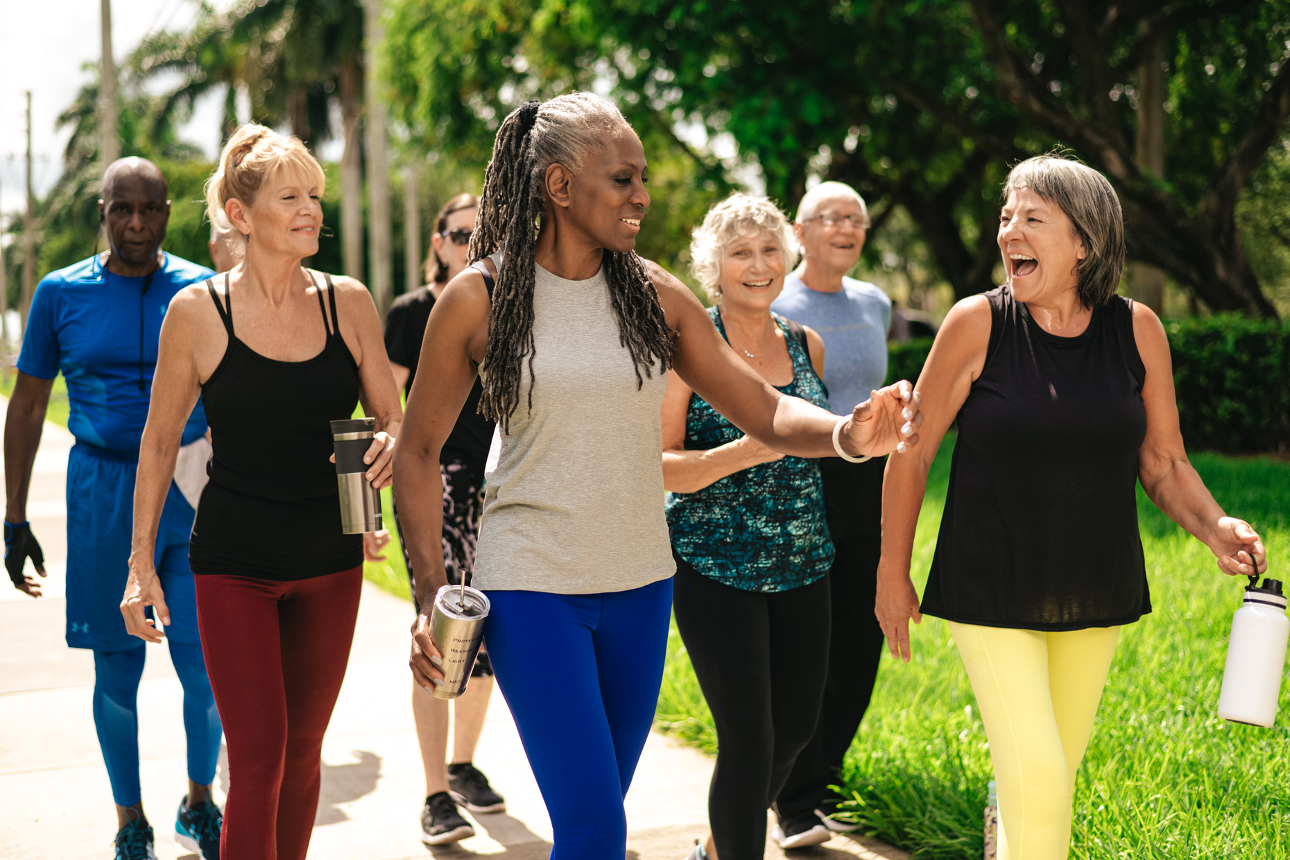 A group of older adults, both men and women, smiling and chatting while walking together outdoors on a sunny day. They are dressed in casual athletic wear, some holding water bottles. The background shows trees and grass, suggesting a park or nature setting—embracing the joy of getting outside every day.