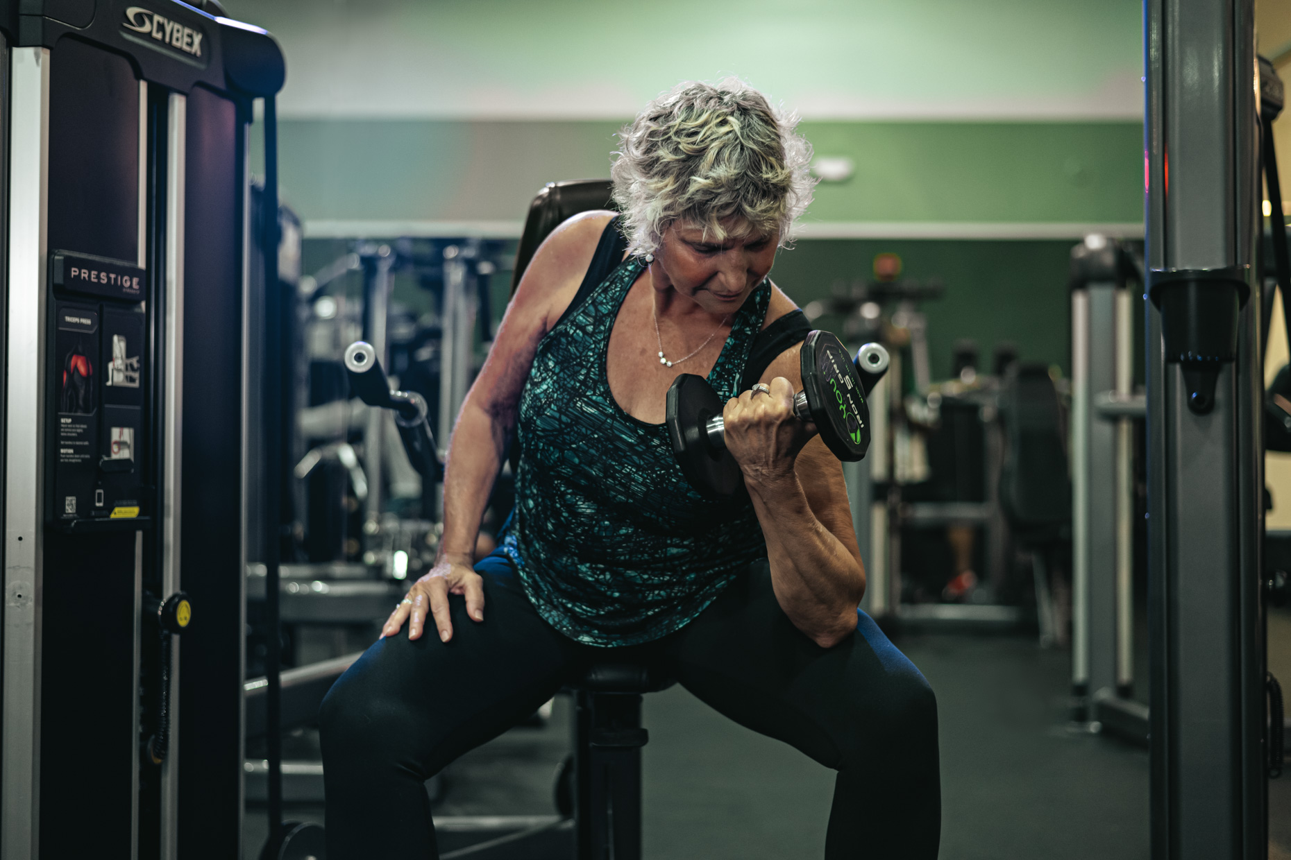 An older woman with short curly hair sits on a bench in a gym, wearing a sleeveless blue top and black leggings, concentrating on her resistance training as she performs a bicep curl with a dumbbell in her right hand. Gym equipment is visible in the background.