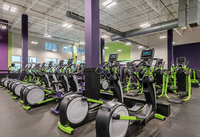 A spacious gym filled with rows of modern elliptical machines and recumbent bikes, featuring vibrant purple and green accents. Bright overhead lights and tall windows illuminate the high-ceiling space, creating an energetic atmosphere for workouts at YouFit Gyms Dania Beach.