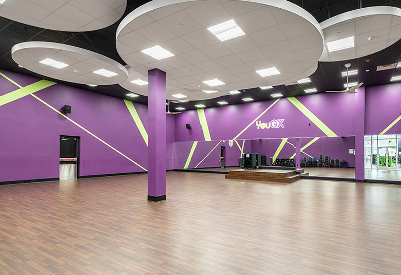 A large, brightly lit dance studio with purple walls, lime green diagonal accents, and a wood floor welcomes you at YouFit Gyms Dania Beach. Circular lighting fixtures adorn the ceiling. A stage with sound equipment is at the front, and chairs are lined up against the walls beneath the logo "YouGX.