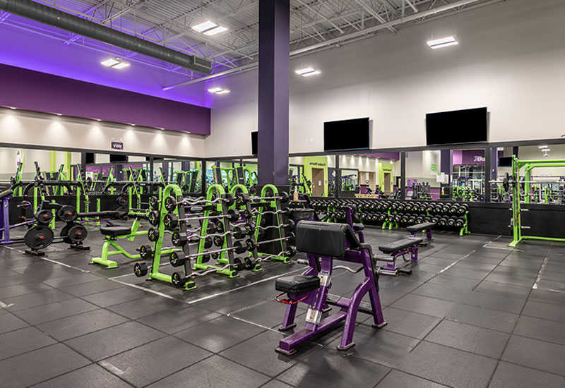 The well-lit interior of YouFit Gyms Dania Beach features a variety of exercise equipment, including weight racks, barbells, benches, and wall mirrors. The equipment is color-coordinated in green and purple, with the floor covered in black rubber tiles.