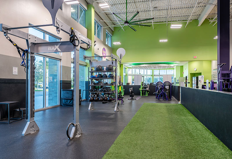 YouFit Gyms Dania Beach features a modern gym with high ceilings, stocked with free weights, exercise machines, and a multi-functional training rig. The facility includes green artificial turf for functional training, large windows for natural light, and fans for ventilation.