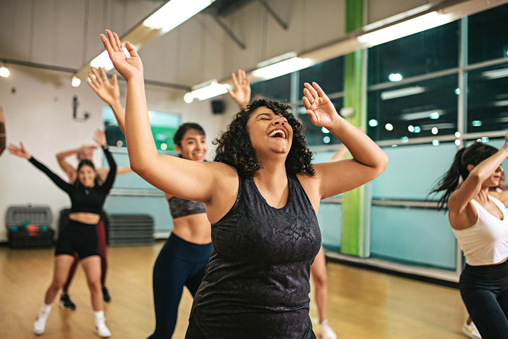 A group of people is energetically dancing in a fitness studio at Youfit Gyms. The focus is on a woman with curly hair, wearing a black tank top, and smiling joyfully with her arms raised. The room has large mirrors and windows, and the atmosphere appears lively and fun.