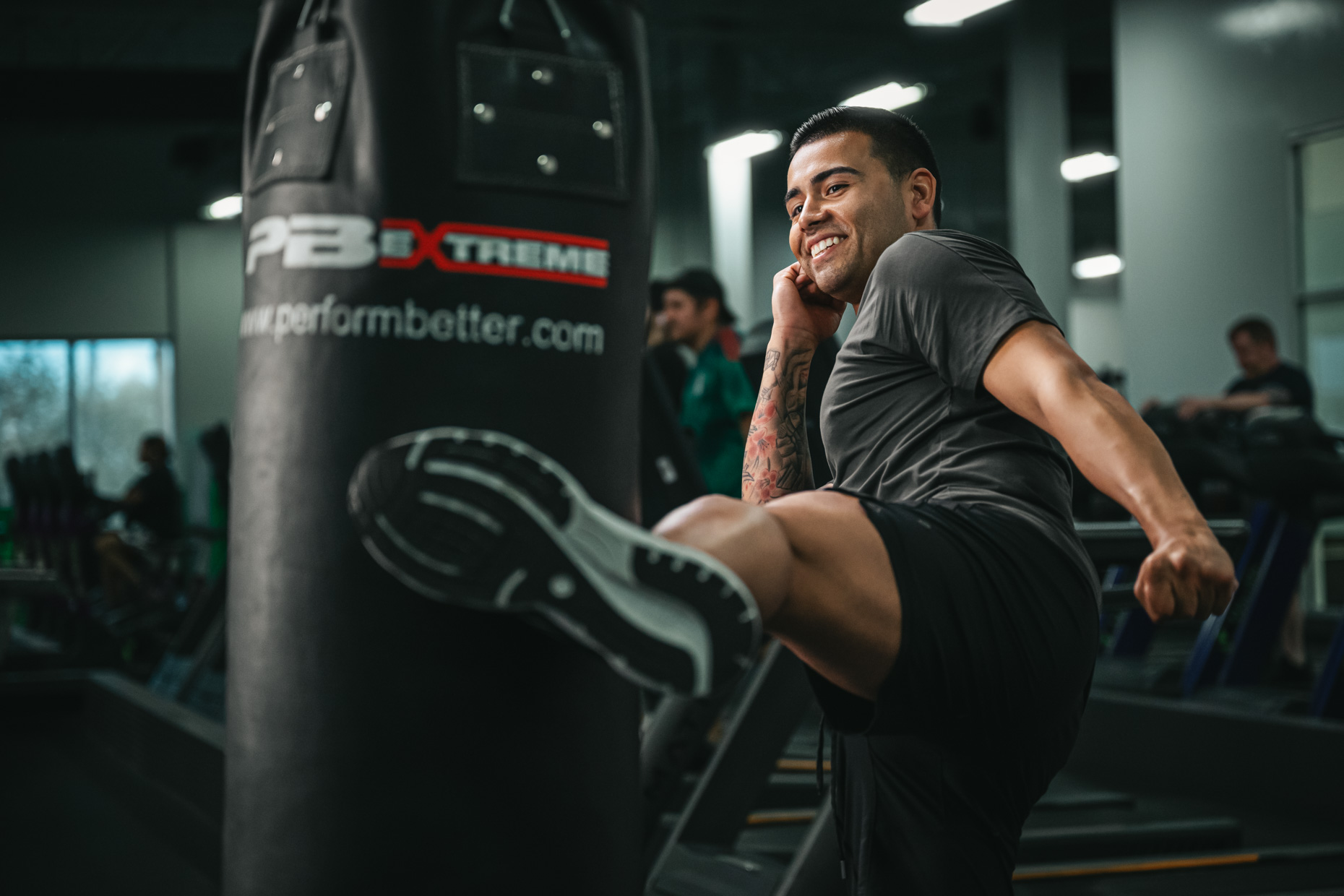 A man in a gym smiles while performing a high kick towards a punching bag. He is wearing a dark athletic shirt and shorts. The background features other gym-goers using exercise equipment, focused on strengthening their joints, and the room is well-lit with fluorescent lights.