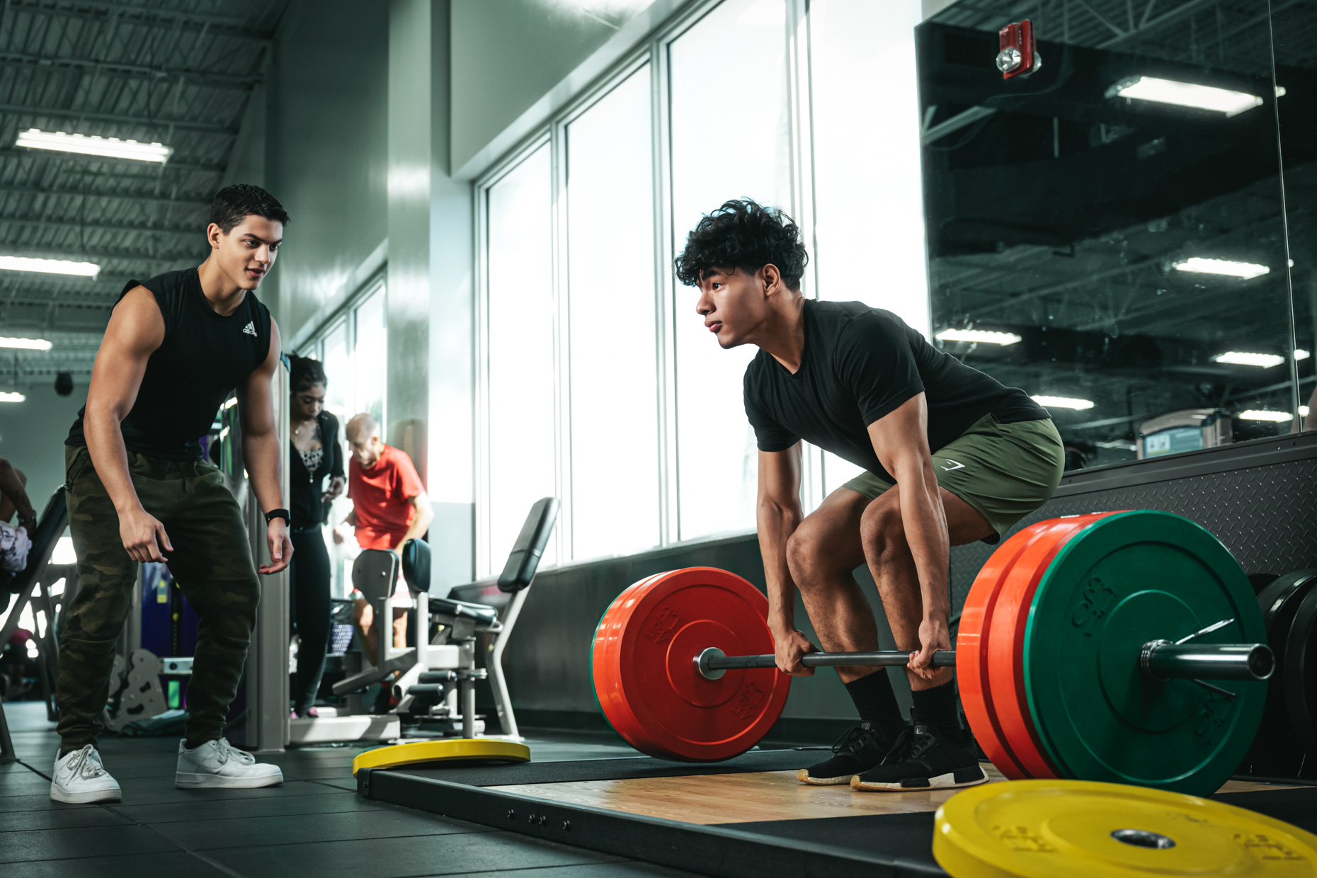 A person stands next to another who is lifting weights at a gym, showcasing sports performance. The person lifting weights is grabbing a barbell with multiple colored weight plates. Both individuals are wearing athletic clothing, and gym equipment and another person can be seen in the background.