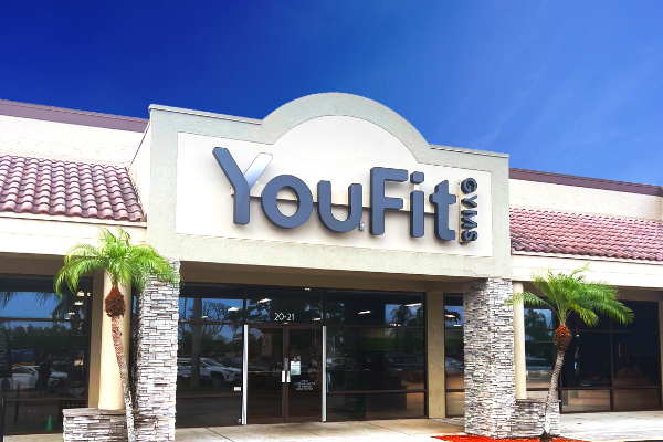 A modern gym storefront with the signage "YouFit" prominently displayed above the entrance. The building features stone columns, large windows, and two small palm trees flanking the entrance—just one of the 5 reasons to join a gym. The sky is clear and blue.