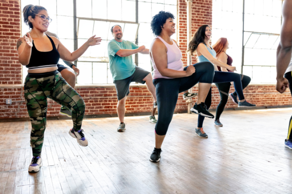 A diverse group of people energetically participate in a fitness class in a room with large windows and exposed brick walls. Mid-motion, lifting one knee while balancing on the other leg, they demonstrate how to add cardio to your workout routine, dressed in various athletic attire.