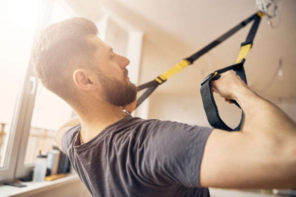 A bearded man in a gray t-shirt is exercising indoors using resistance straps attached to the ceiling. He is focused and appears to be pulling the straps towards himself as part of his gym workout routine for busy dads. Sunlight streams in through a window in the background.