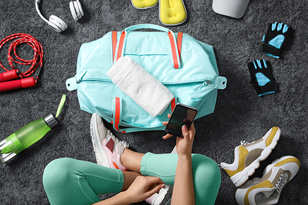 A person sitting on a gray carpet is holding a smartphone. Next to them is a blue gym bag with a white towel on top. Surrounding items include sneakers, a jump rope, headphones, a water bottle, weightlifting gloves, and yellow slides—everything you need for the perfect gym starter kit.