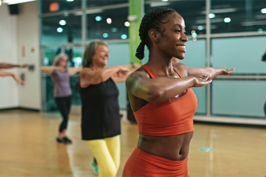 A group of people participate in an indoor fitness class at Youfit Gyms. The focus is on a person in the foreground, wearing a red sports outfit, smiling and extending their arms. Other participants in colorful athletic clothing follow along. The background is a spacious gym.