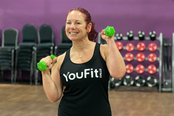 A woman with red hair smiles while lifting green dumbbells. She is wearing a black tank top with the words "YouFit Gyms" printed on it, proud to be part of the SilverSneakers program. The background shows stacked chairs and a rack of weighted balls against a purple wall.