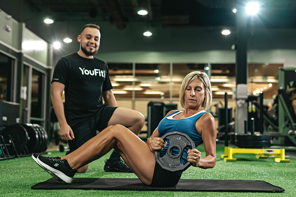 A woman performs a seated Russian twist holding a weight plate while sitting on an exercise mat in a gym. A man, who aspires to become a personal trainer, stands nearby, smiling and watching her workout. The gym has various fitness equipment in the background and bright lights overhead.