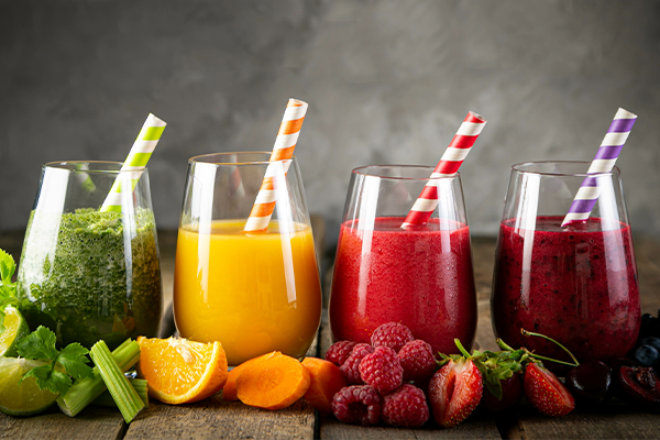 Four glasses of colorful smoothies to help prevent bloating sit on a wooden table, each with a striped straw. From left to right, the smoothies are green, yellow, red, and purple. Various fruits and vegetables such as celery, orange slices, raspberries, and strawberries are placed in front.