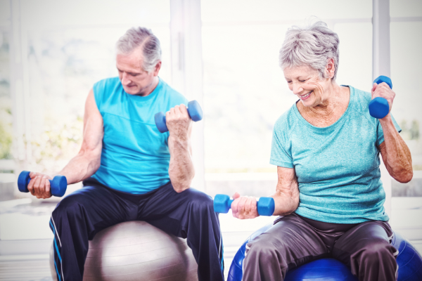 An elderly man and woman are seated on exercise balls, lifting blue dumbbells. Both are wearing light blue workout attire and smiling, seemingly enjoying their weight workouts for seniors. The background is bright with large windows letting in natural light.