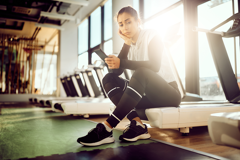 A young woman in athletic wear sits on a gym bench, glancing at her phone with a bored expression. Several treadmills are lined up in the background as sunlight streams through large windows, illuminating the space where exercise and maintaining a healthy lifestyle take center stage.