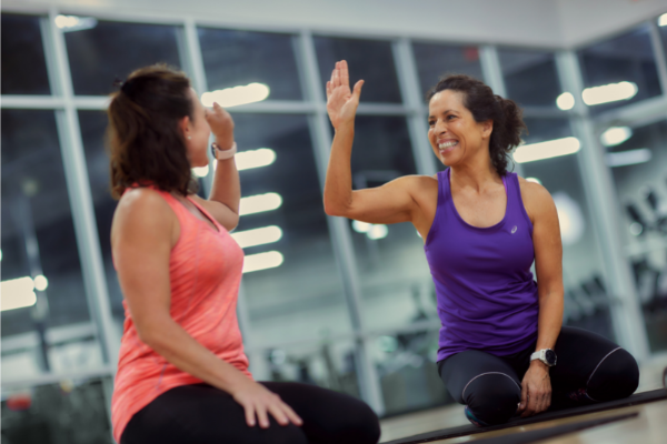 Two women in athletic wear sit on a gym floor, facing each other and giving a high-five after a successful session. They are smiling and appear to be in a positive, upbeat mood. It's clear that working out with a friend makes exercise more enjoyable. In the background, large windows blur the view of gym equipment.