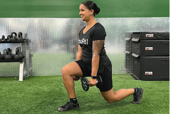 A woman in athletic wear performs a lunge exercise in a gym, holding a weight in each hand. She is smiling, with gym equipment in the background, including kettlebells and stacked boxes. The wall is covered with silver diamond plate panels—a perfect scene for workouts to help you break a sweat.