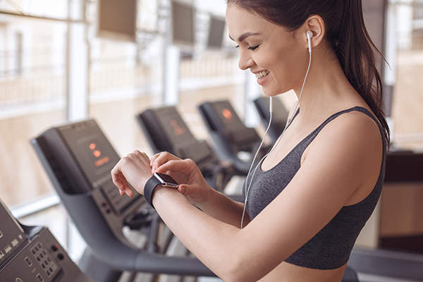 busy person woman stands in a gym with treadmills in the background, wearing workout attire and earphones. She smiles while looking at her smartwatch, ready to get the most out of the YouFit Gyms app.