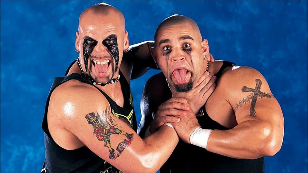 Two people pose playfully against a blue background, with exaggerated facial expressions and tongues out. Sporting black clothing and arms inked like Glenn Ruth's, one playfully holds the other's neck in a mock wrestling grip.